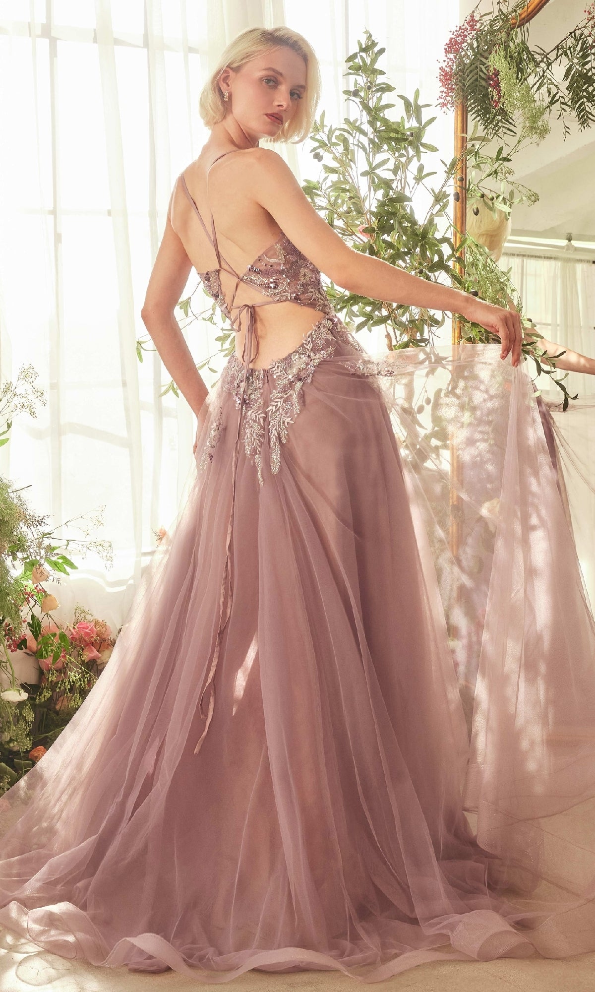 A woman stands in a sunlit room surrounded by flowers, wearing the Andrea & Leo A1419 Violet Purple Long Prom Dress with a beaded bodice, open back, lace-up detail, and floral embroidery.